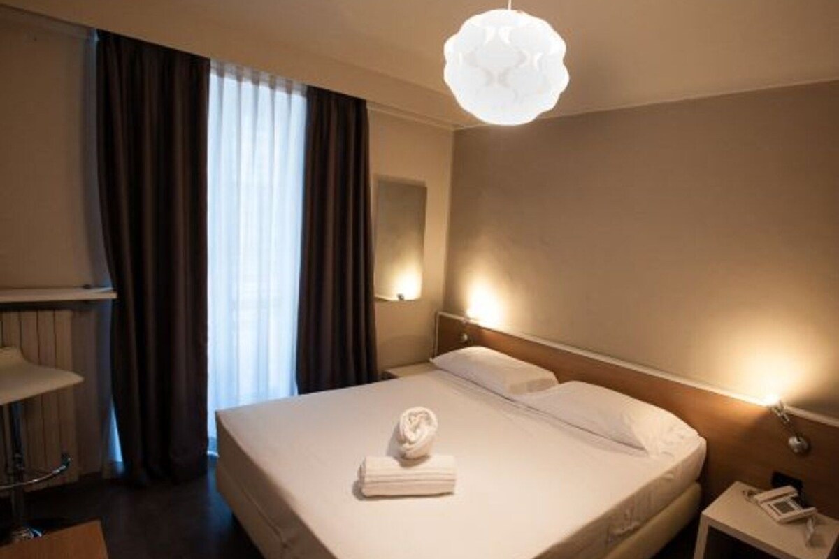 A hotel room with a large bed, a bedside table with a lamp, a wall-mounted light, and a window with curtains.