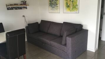 En moderne stue med en grå sofa, en sort lænestol og et lille bord. To billeder hænger på væggen, og der står en potteplante på en hylde.