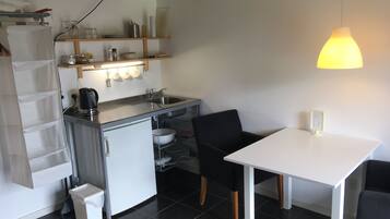 A compact kitchen with a sink, a kettle, and a small refrigerator. There is a white table with two chairs and a hanging light fixture above.