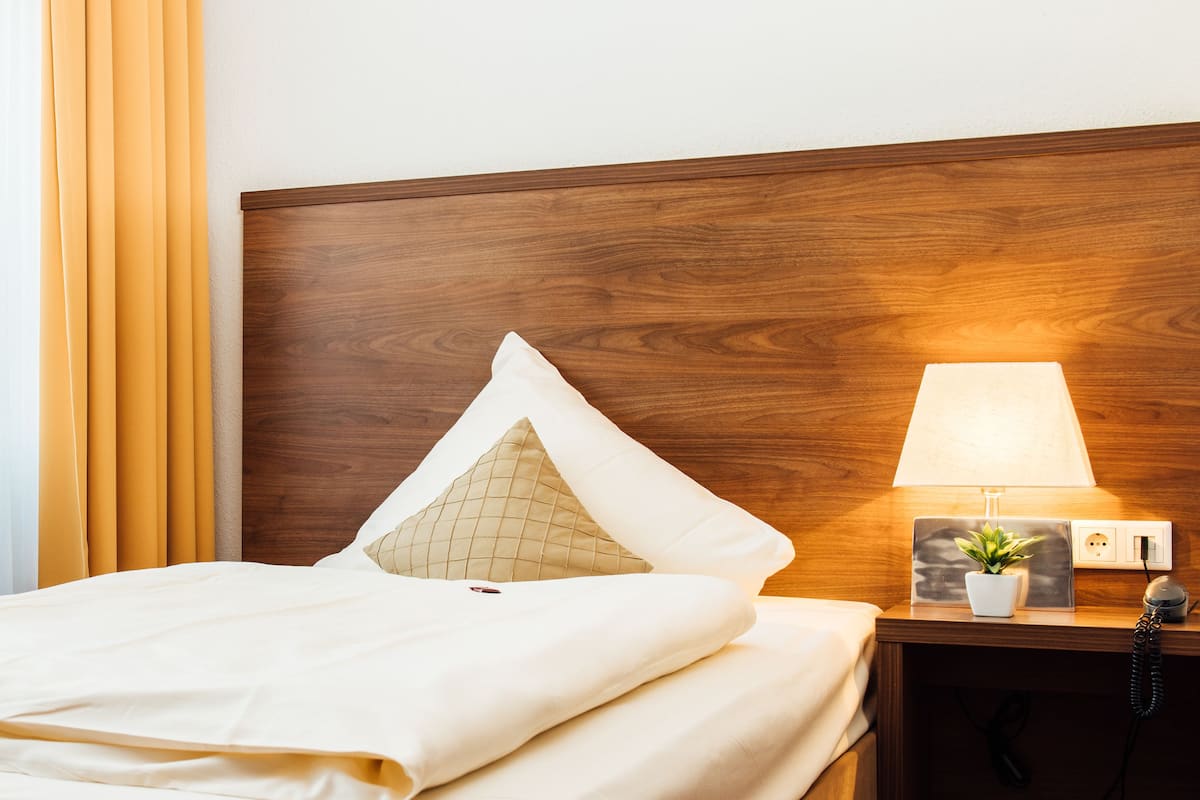 A neatly made bed with white linens, a wooden headboard, a bedside lamp, a small potted plant, and a nightstand with a telephone.