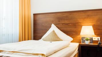 A neatly made bed with white linens, a wooden headboard, a bedside lamp, a small potted plant, and a nightstand with a telephone.