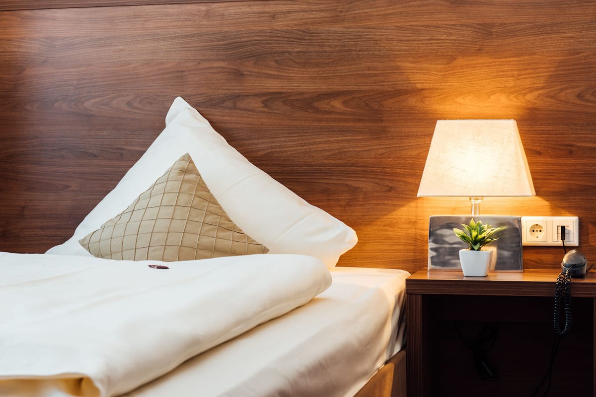 A neatly made bed with a white comforter and two pillows, a bedside table with a lamp, a small potted plant, and a power outlet.