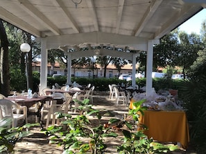 An outdoor covered patio area with tables and chairs, surrounded by greenery and buildings in the background. - Corallo Country Hotel & SPA (Noci)