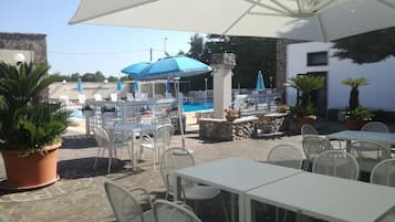 An outdoor patio with white tables and chairs, a pool area with blue umbrellas, and potted plants.