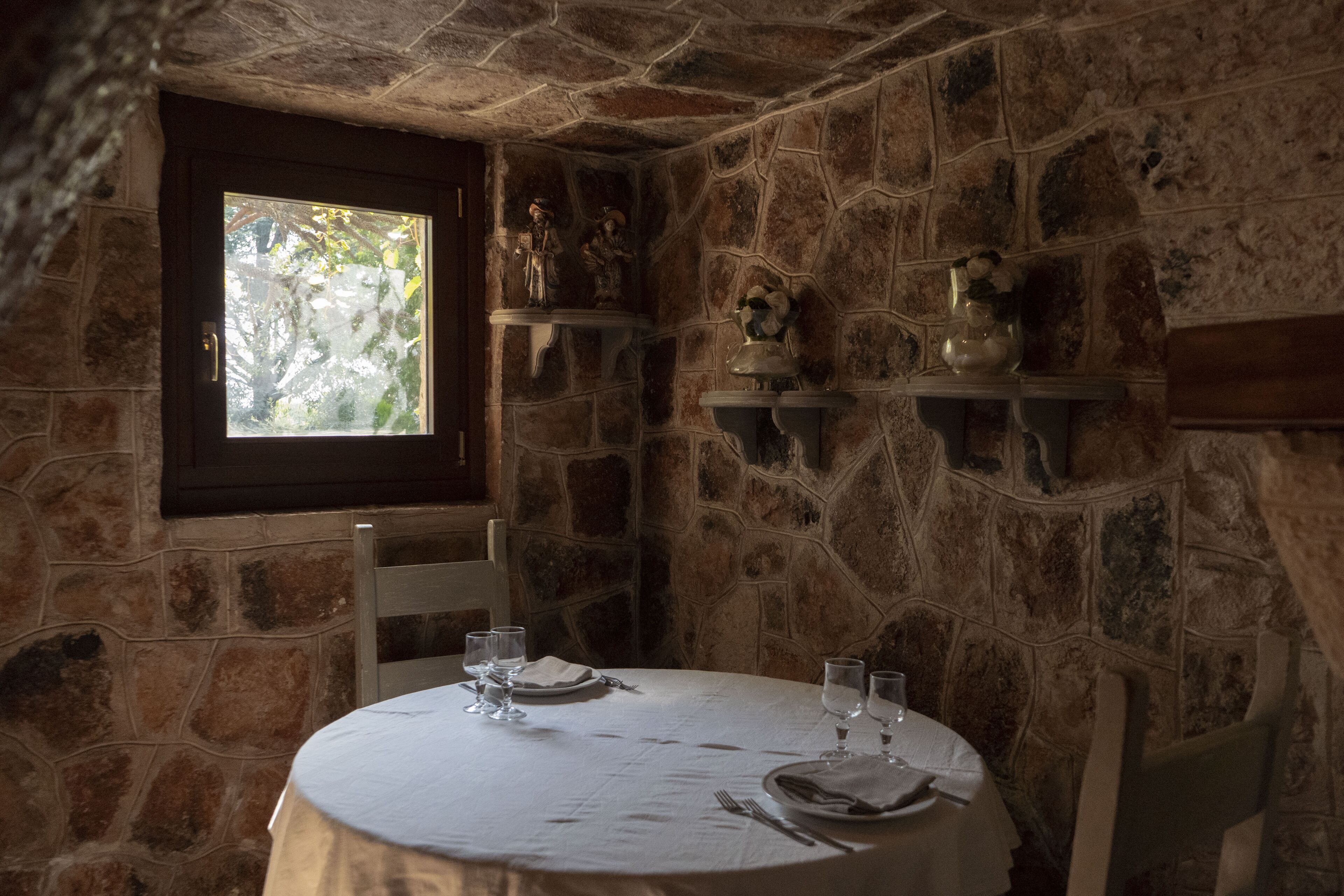 Un'area ristorazione con pareti in pietra, un tavolo rotondo apparecchiato per due persone, una finestra con vista sugli alberi e oggetti decorativi sugli scaffali.
