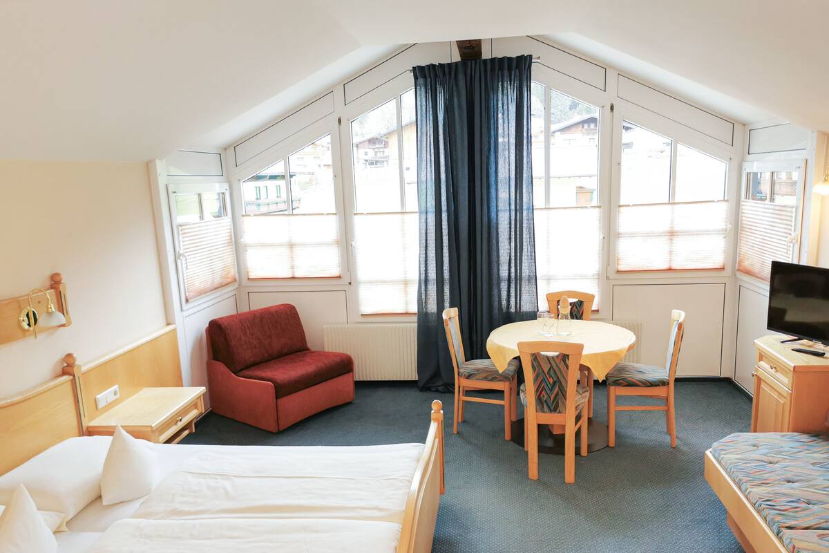 Une chambre d’hôtel avec un lit, un fauteuil rouge, une table à manger avec des chaises, une télévision et de grandes fenêtres avec des rideaux.