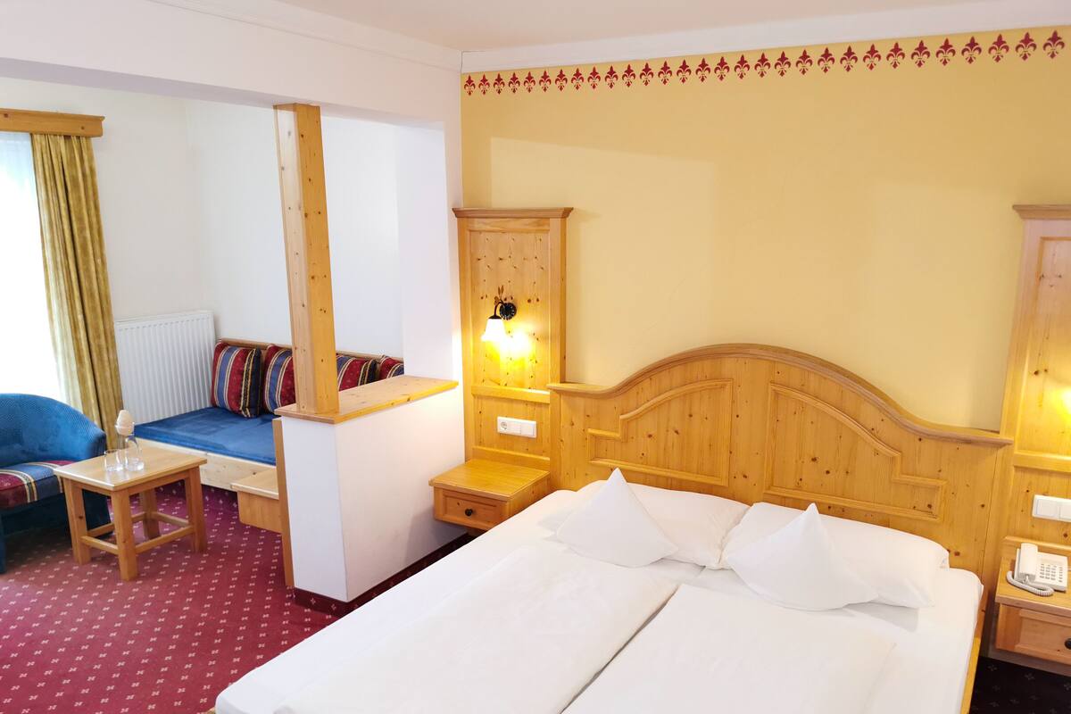 Une chambre d’hôtel avec un grand lit, une tête de lit en bois, une table de chevet avec une lampe, une petite armoire en bois et un canapé agrémenté de coussins colorés.
