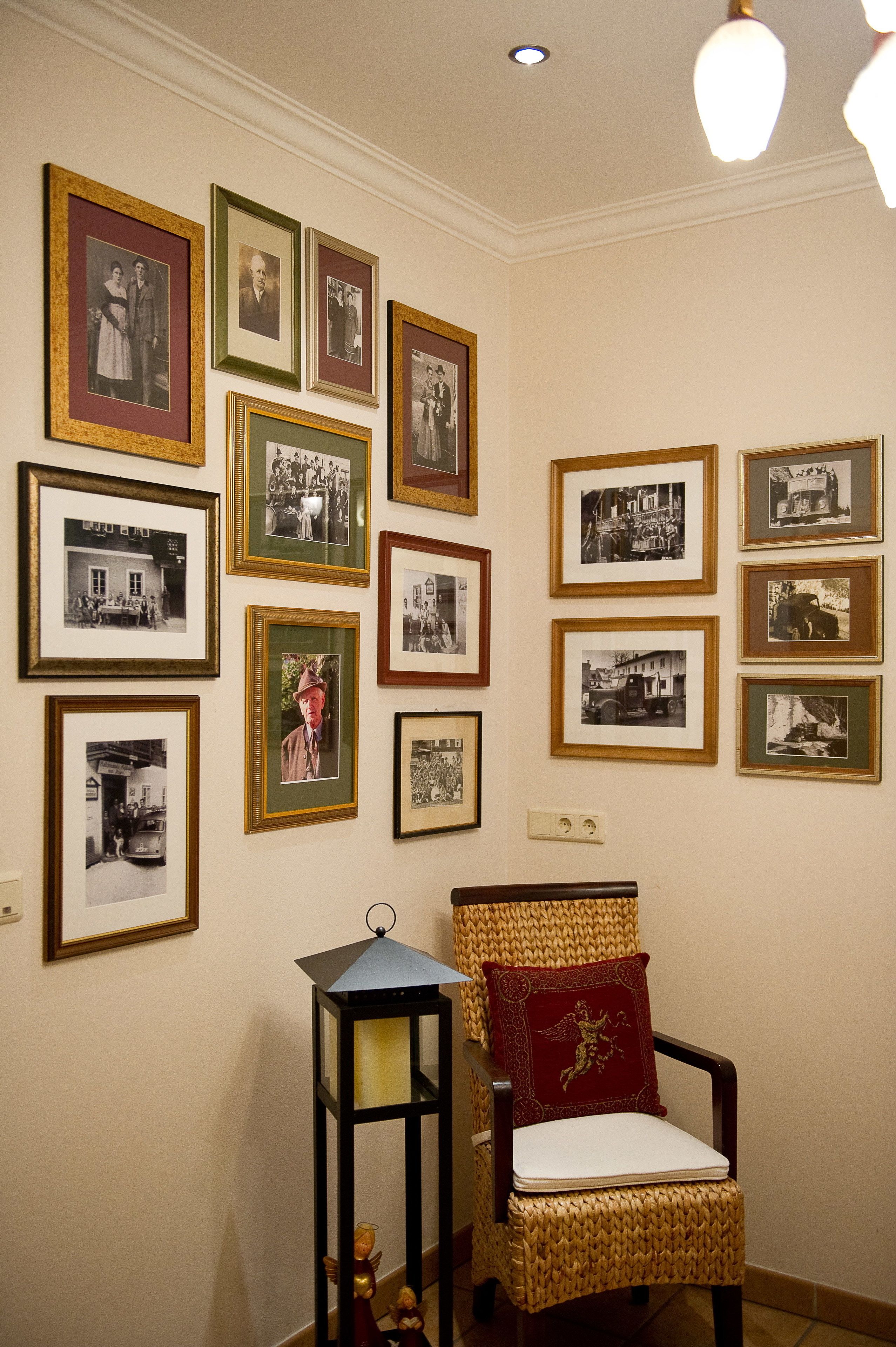 Ein Zimmer mit einer Wand, die mit gerahmten Schwarz-Weiß-Fotos geschmückt ist, ein Korbstuhl mit rotem Kissen und ein schwarzer Laternenleuchter.