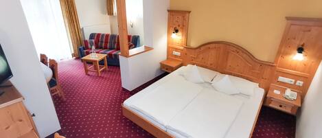 Quarto de hotel com uma cama grande, cabeceira de madeira, uma mesinha com uma cadeira, um sofá e uma televisão.