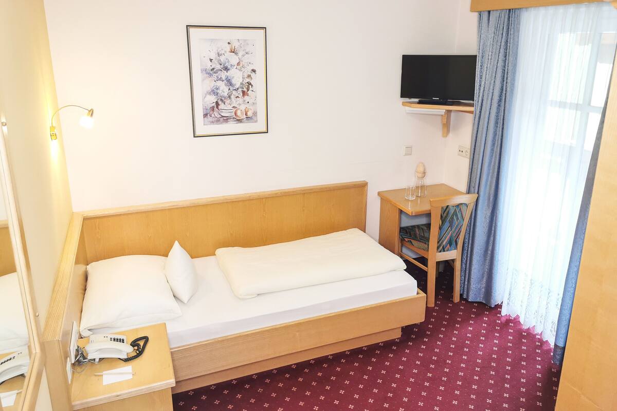 Une chambre d’hôtel avec un lit en bois, une table de chevet avec un téléphone et des papiers, une petite table avec un verre, un téléviseur fixé au mur et une fenêtre avec des rideaux.