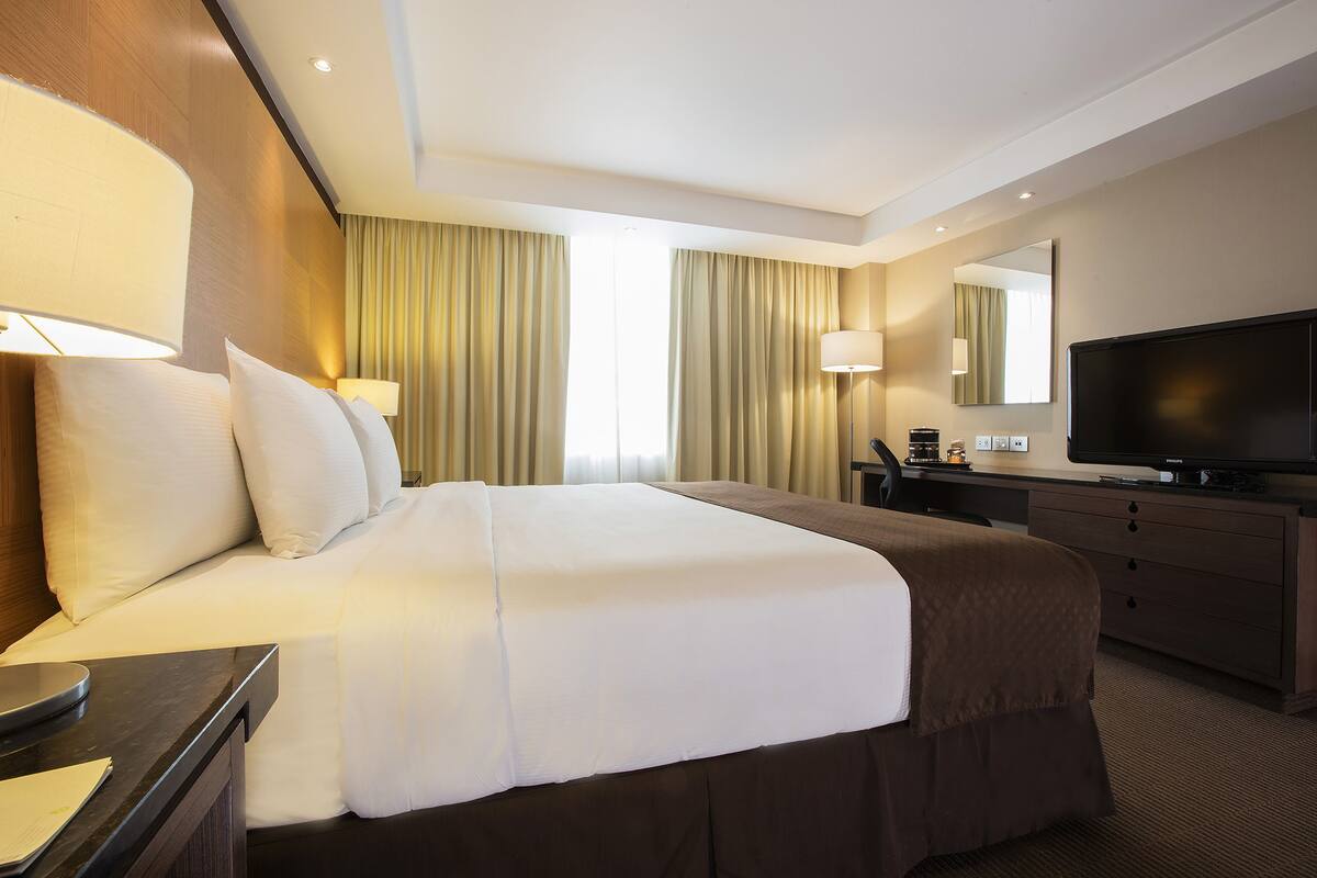 A hotel room with a large bed, a flat-screen TV, a desk with a lamp, and a dresser.