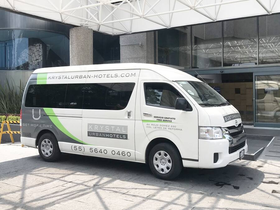 A white van with "KRYSTALURBAN-HOTELS.COM" and "SERVICIO GRATUITO FREE SERVICE" on the side, parked in front of a modern building with large windows and a geometric glass facade.