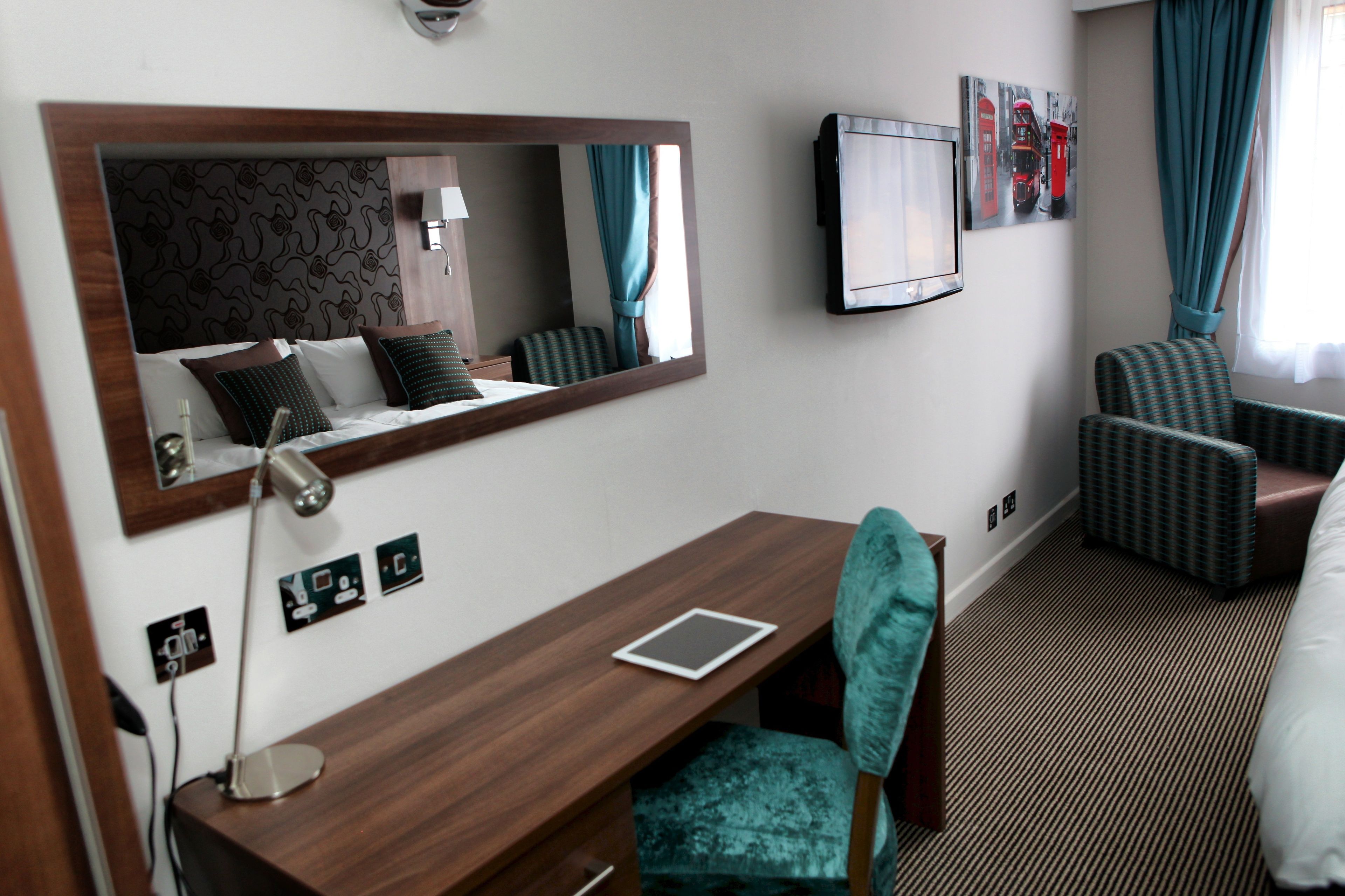 A hotel room with a wooden desk, a mirror, a television, a patterned chair, and a bed.
