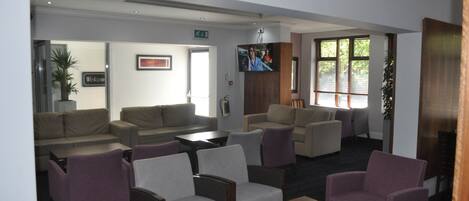 A hotel lobby with sofas, armchairs, a coffee table, a TV mounted on the wall, and a 'Welcome' sign.