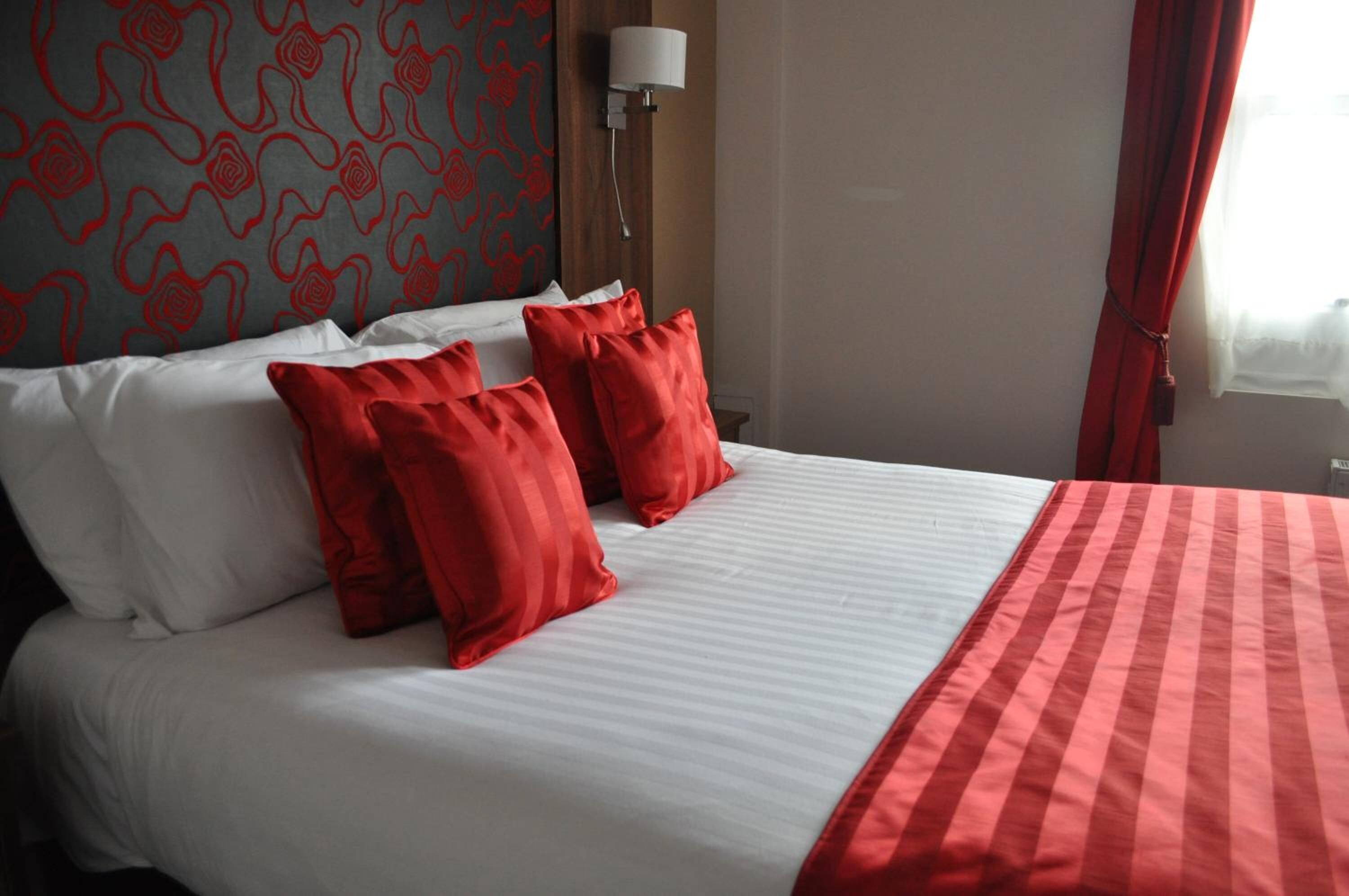 Habitación de hotel con cama, almohadas rojas y blancas, manta a rayas rojas y blancas, pared estampada y ventana con cortinas rojas.