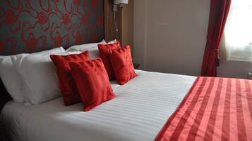 Habitación de hotel con cama, almohadas rojas y blancas, manta a rayas rojas y blancas, pared estampada y ventana con cortinas rojas.