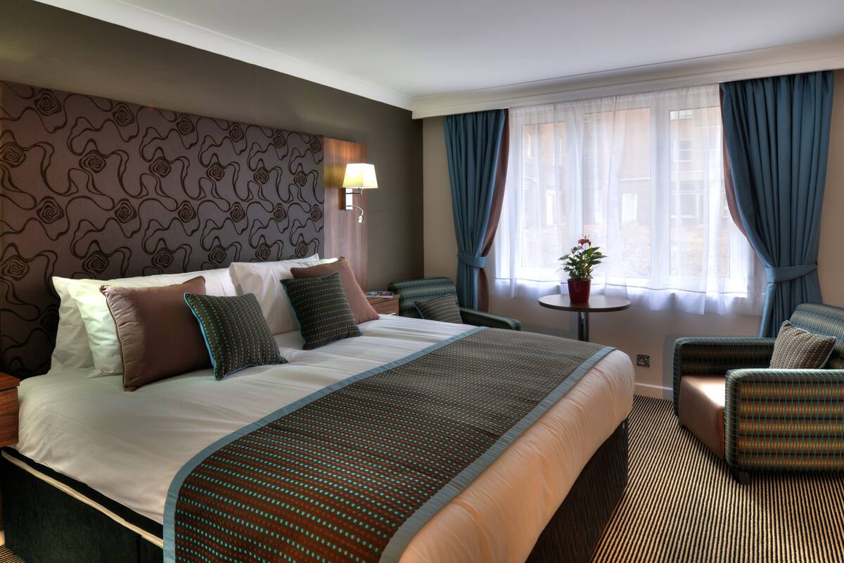 Une chambre d’hôtel avec un grand lit, un coin salon comprenant une chaise et une petite table, et une fenêtre avec des rideaux.