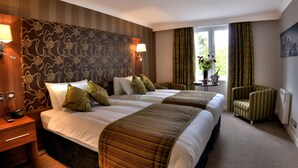 A hotel room with two beds, a small table with a vase of flowers, a chair, and a lamp on the wall.