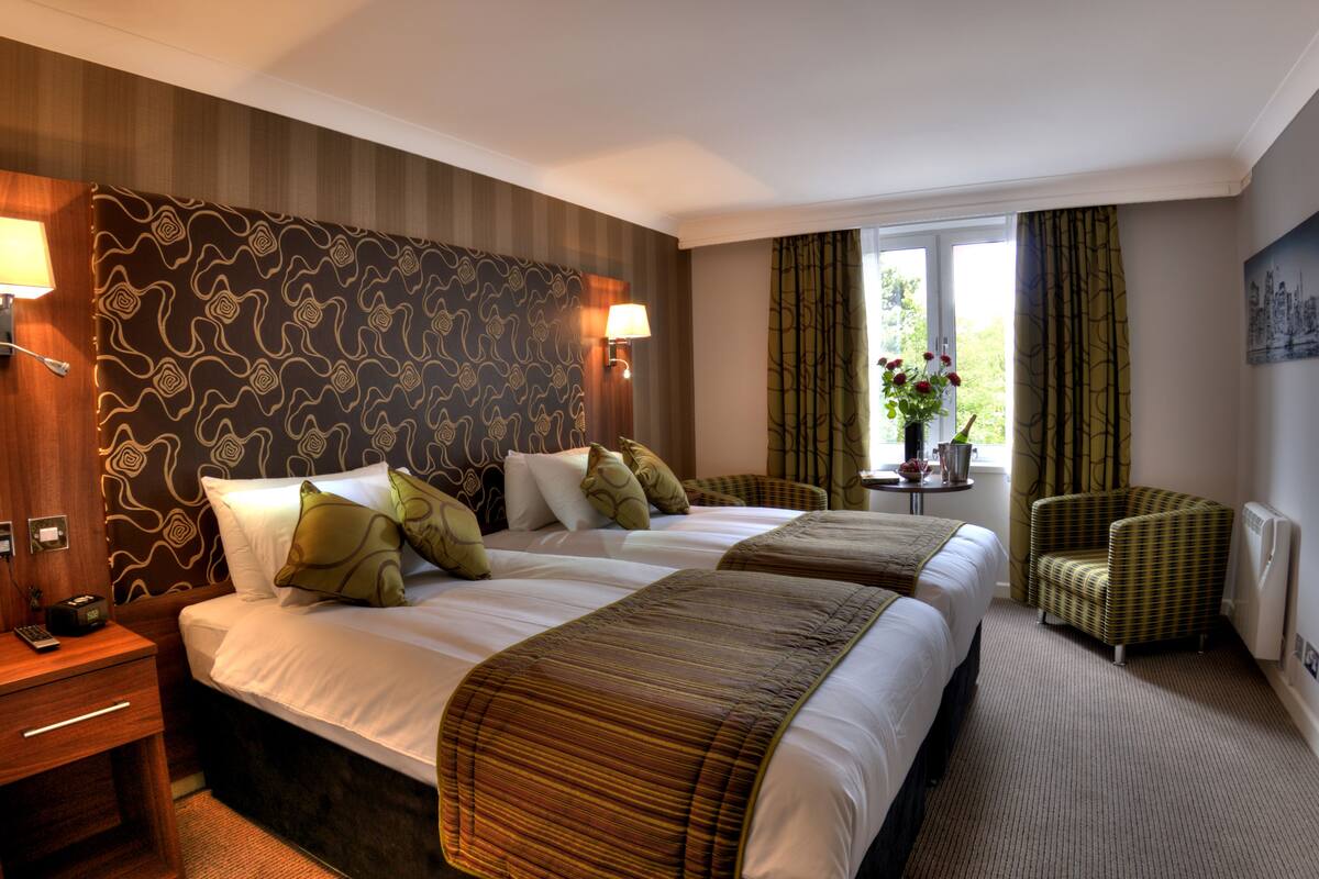 Une chambre d’hôtel avec deux lits, une petite table ornée d’un vase de fleurs, une chaise et une lampe fixée au mur.