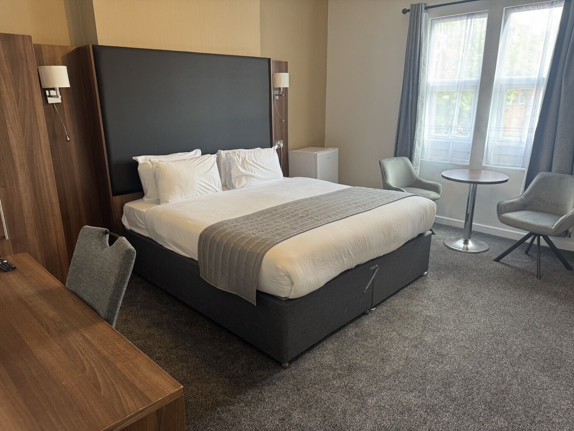 A hotel room with a large bed, a desk, a chair, a small table, and a window with curtains.