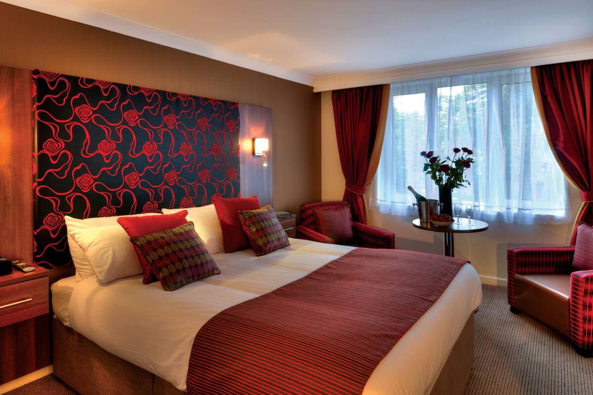 Une chambre d’hôtel avec un grand lit, une literie rouge et blanche, une tête de lit rouge à motifs, une petite table avec un vase de fleurs et des rideaux rouges.