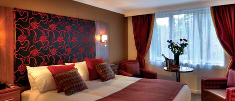 Une chambre d’hôtel avec un grand lit, une literie rouge et blanche, une tête de lit rouge à motifs, une petite table avec un vase de fleurs et des rideaux rouges.