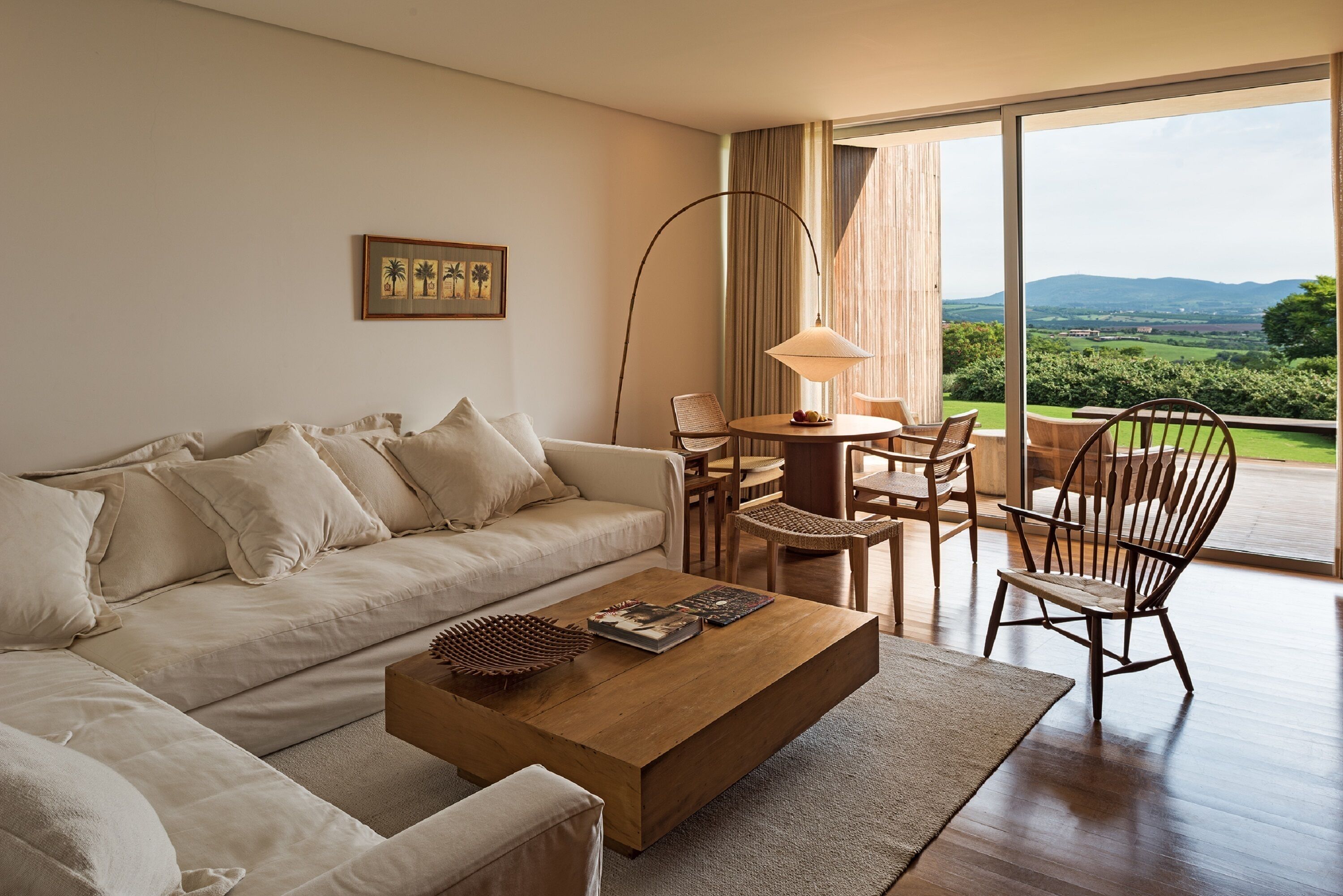 A modern living room with a sofa, coffee table, and chairs, a large floor lamp, and a view of greenery through large windows.