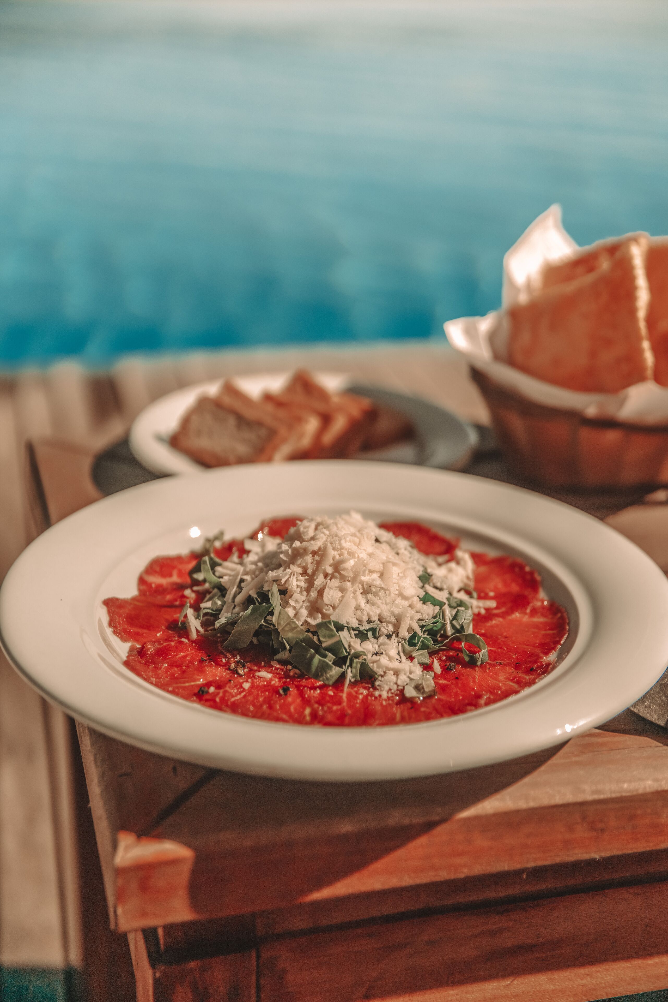 Ein Teller Essen mit roter Soße, Grünzeug und Käse, dazu ein Brotkorb auf einem Holztisch in Poolnähe.
