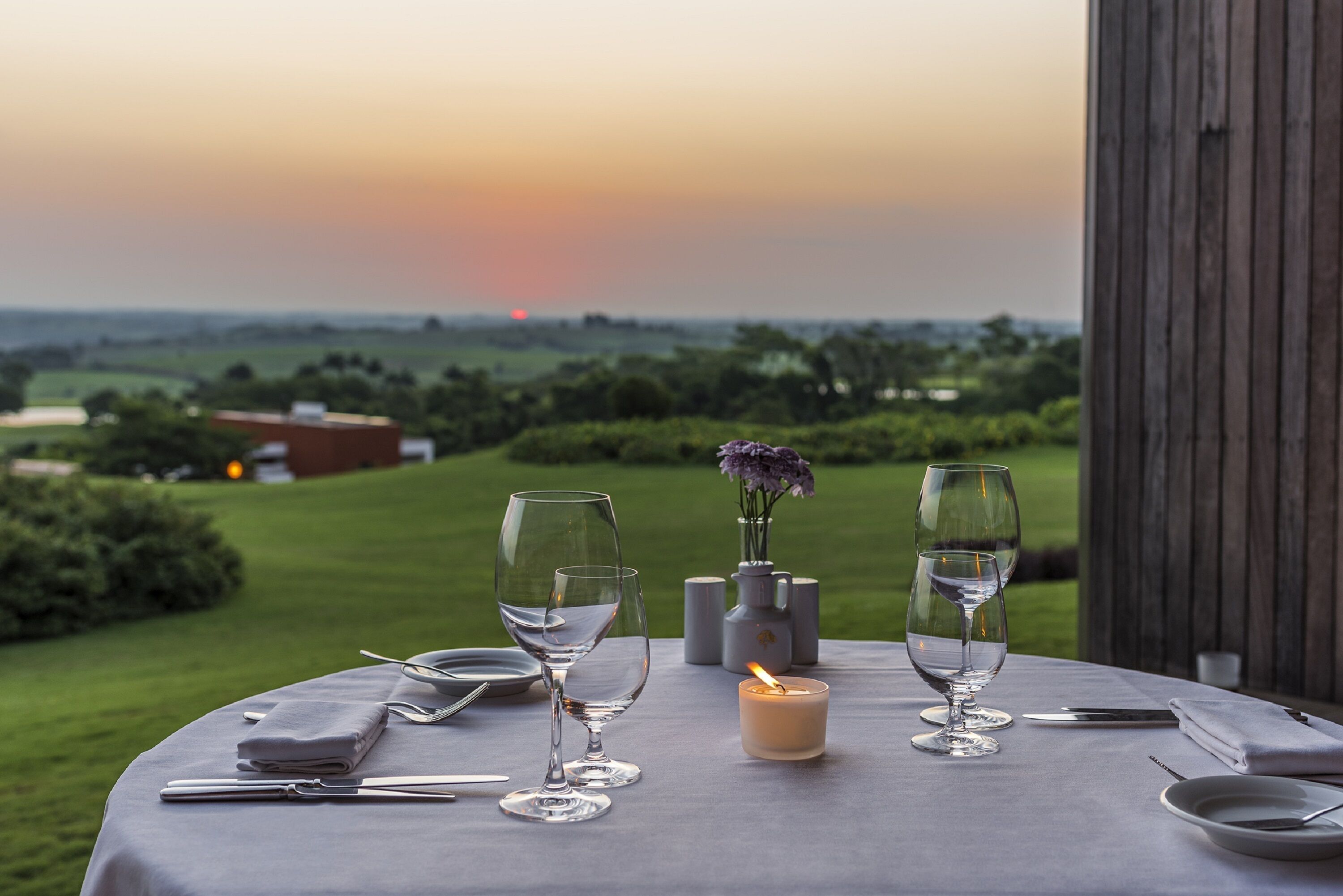 Une table dressée pour deux personnes, avec des verres à vin, une bougie et un vase rempli de fleurs, donnant sur un paysage magnifique.