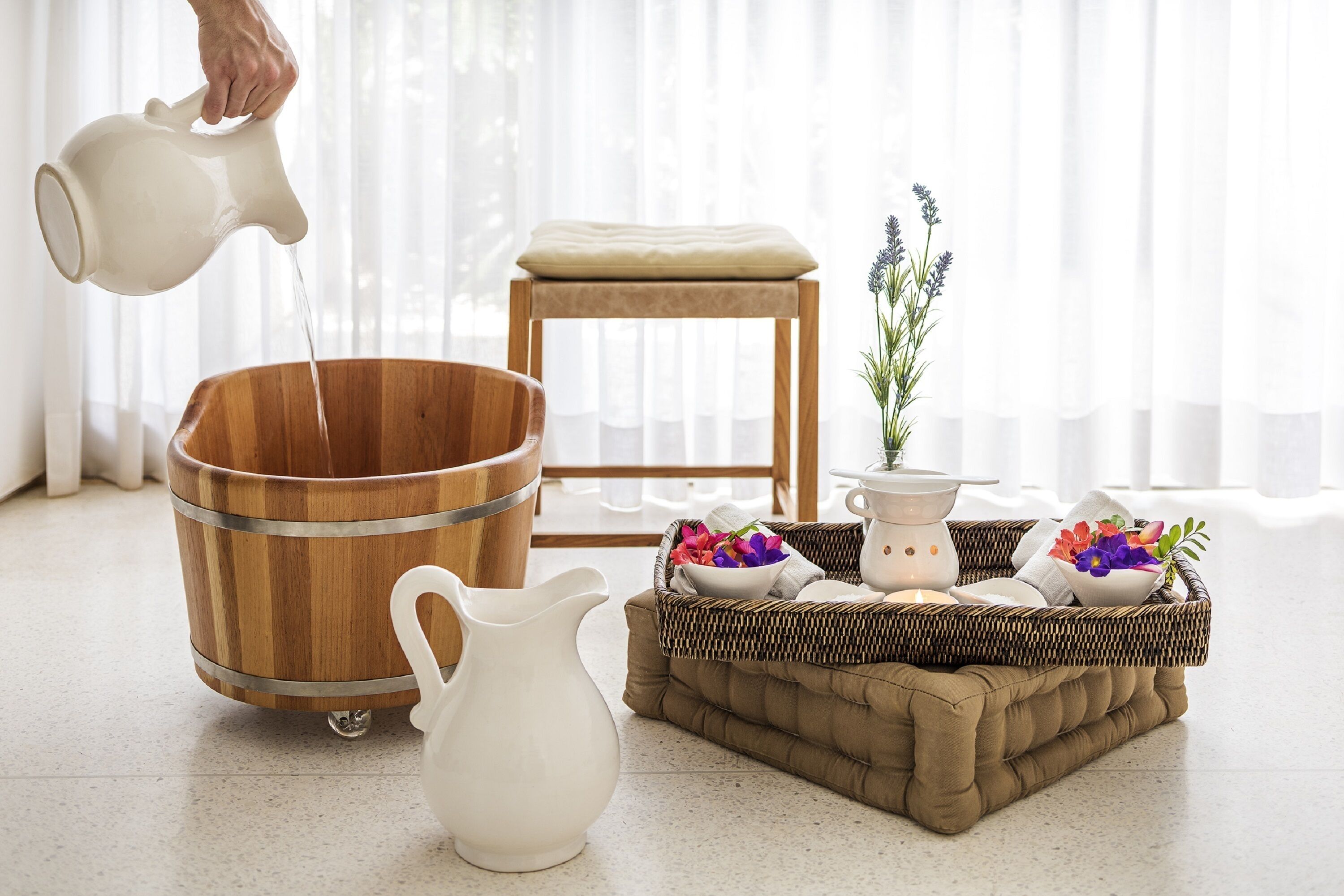 Un balde de madera, una jarra blanca y una cesta de mimbre con flores y una vela sobre un taburete acolchado.