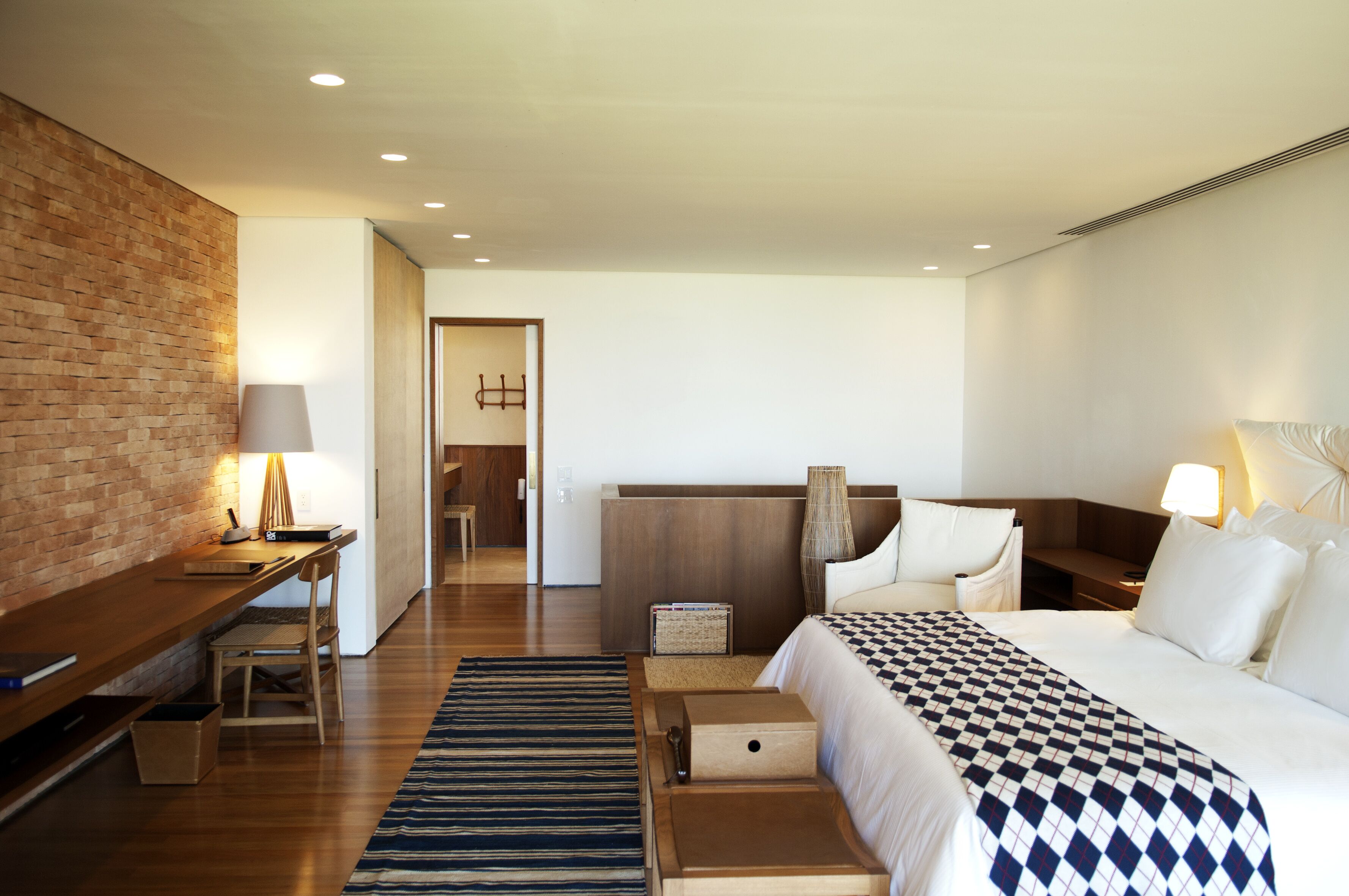 A hotel room with a wooden desk, a bed with a checkered bedspread, a nightstand, a lamp, and a brick accent wall.