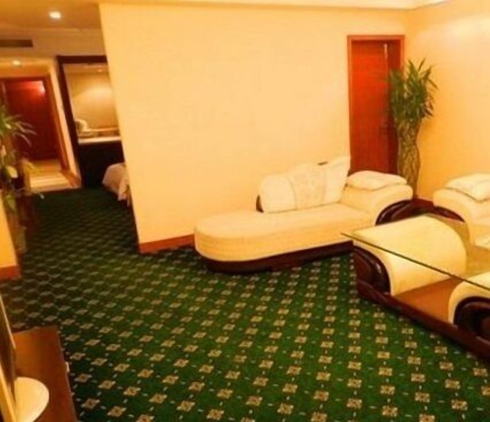 Een hotelkamer met een groen tapijt met patroon, een witte bank, een glazen salontafel en een houten bijzettafeltje.