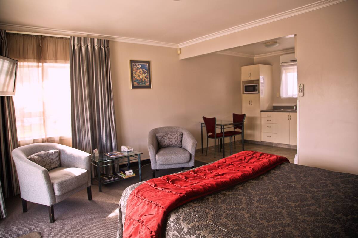 Habitación de hotel con cama, una silla, una mesita con lámpara, un escritorio con silla y una kitchenette al fondo.