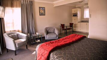 A hotel room with a bed, a chair, a small table with a lamp, a desk with a chair, and a kitchenette in the background.