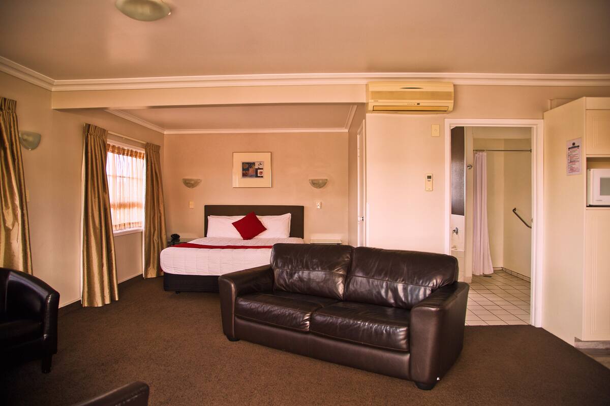 Habitación de hotel con cama, sofá de cuero, una silla y un baño visible a través de una puerta abierta.