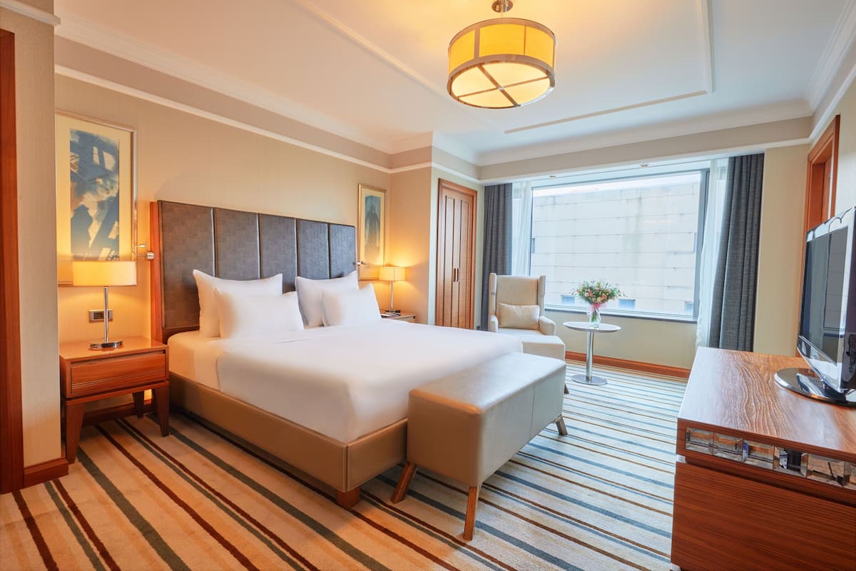 A hotel room with a large bed, a desk with a flat-screen TV, a small round table with a vase, and a window with curtains.