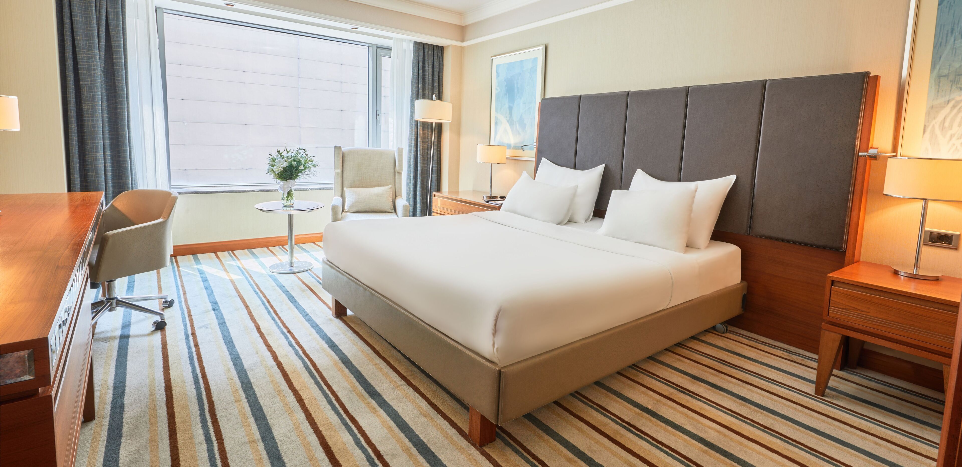 Une chambre d’hôtel avec un grand lit, un bureau, une chaise, une petite table avec un vase et une fenêtre avec des rideaux.