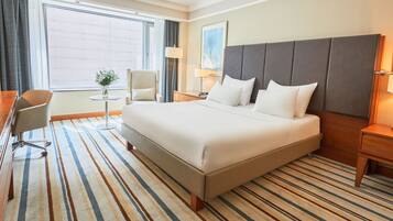 Une chambre d’hôtel avec un grand lit, un bureau, une chaise, une petite table avec un vase et une fenêtre avec des rideaux.