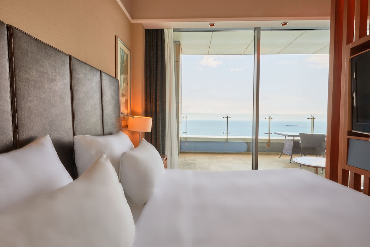 A hotel room with a large bed, a view of the sea, and a balcony with a table and chairs.