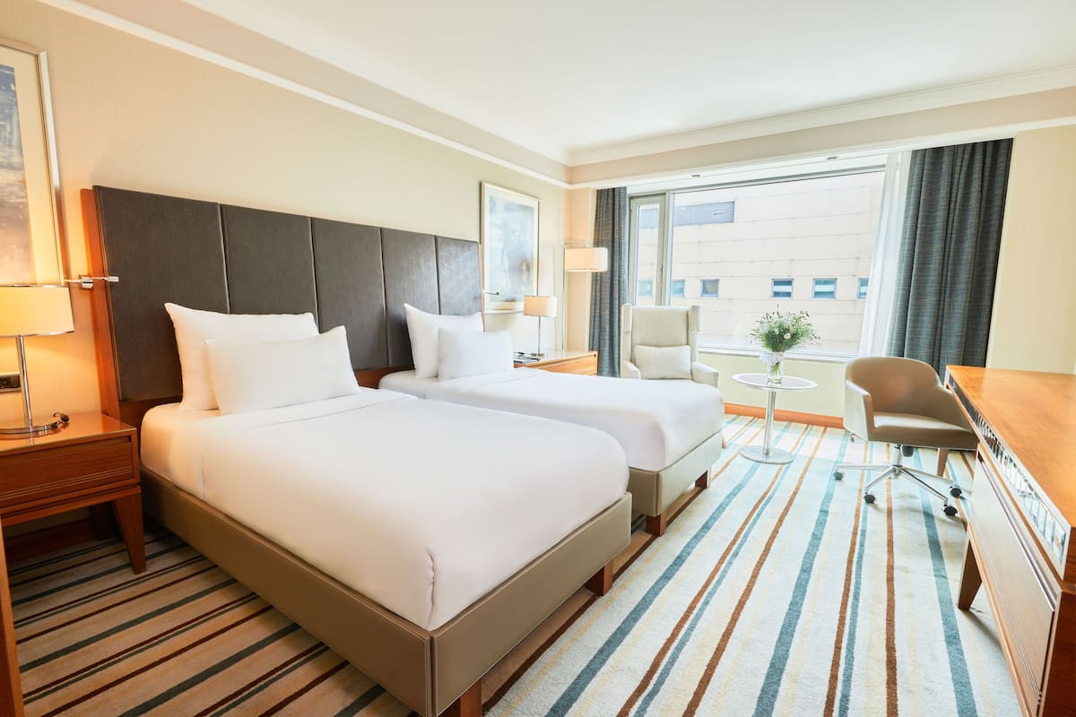 A hotel room with two beds, a desk, a chair, a lamp, and a window with curtains.