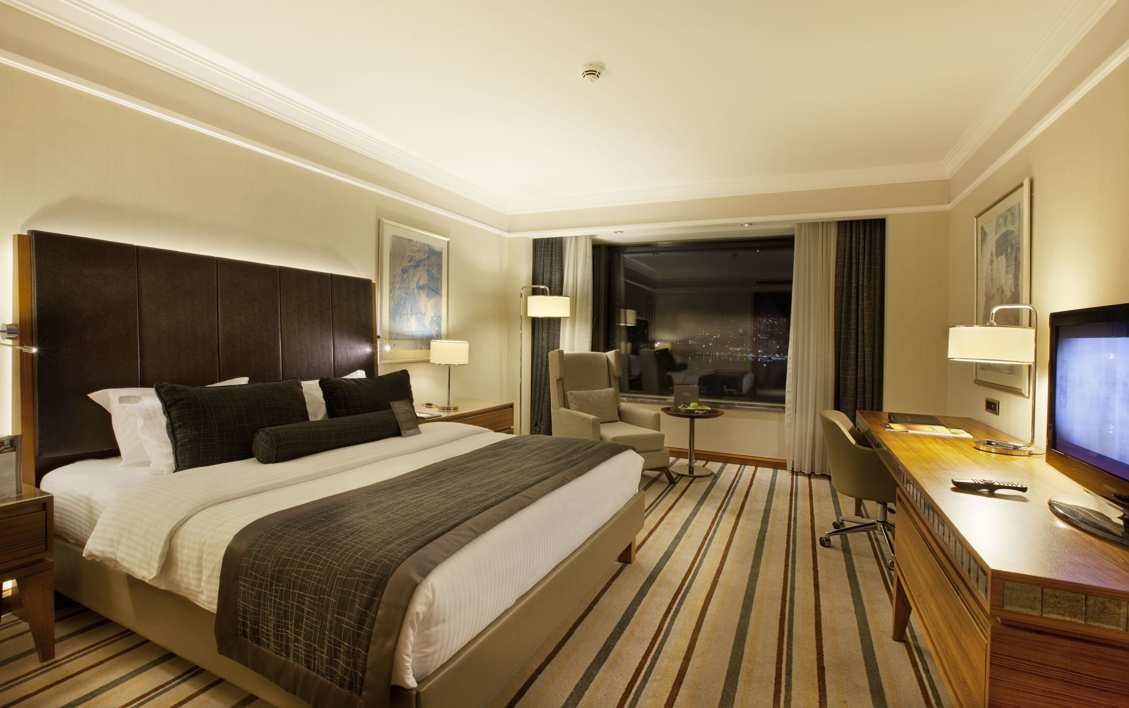 Habitación de hotel con una cama grande, un escritorio con computadora, una silla, un televisor y una ventana con vista a la ciudad.