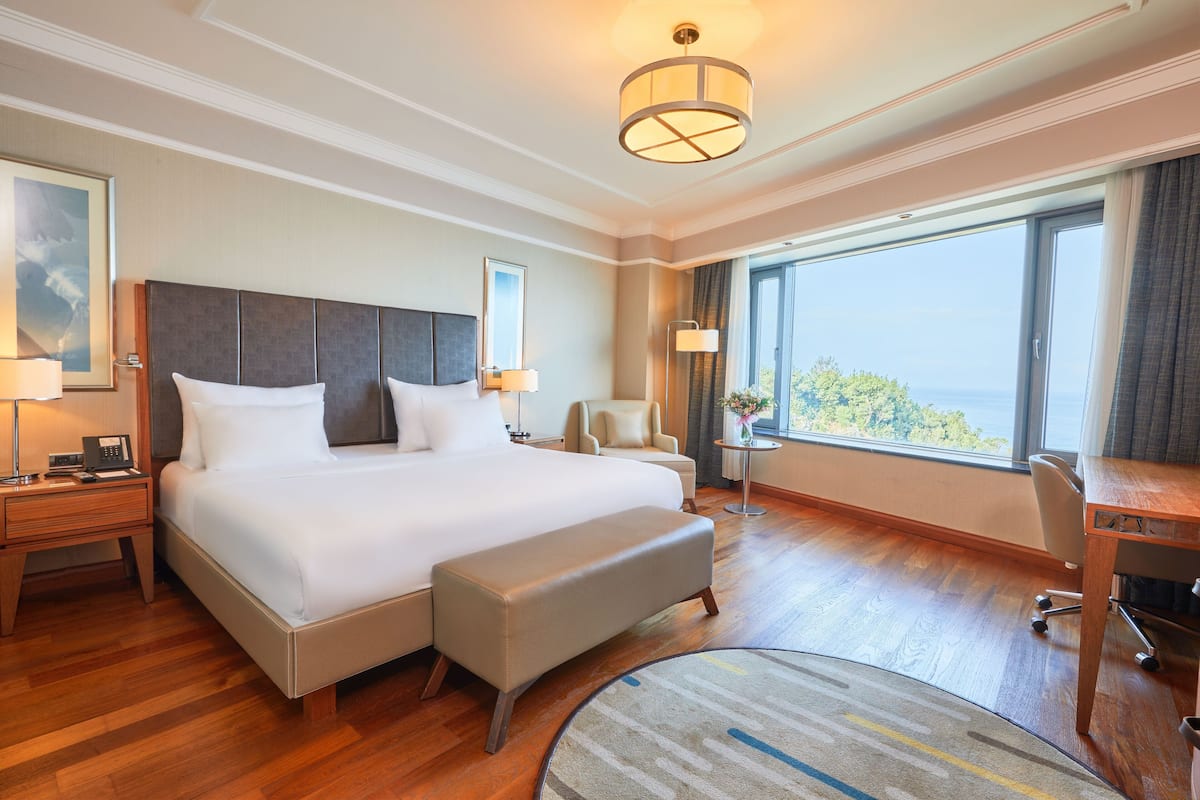 A hotel room with a large bed, a desk, a chair, a small table, a bedside lamp, a nightstand, and a view of the outdoors.