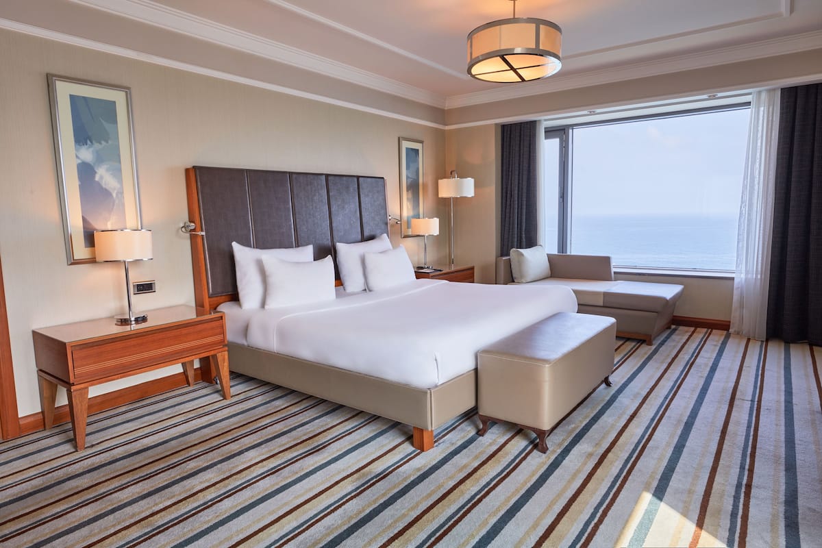 A hotel room with a large bed, a bedside table, a chair, a small table, and a large window with a view of the ocean.
