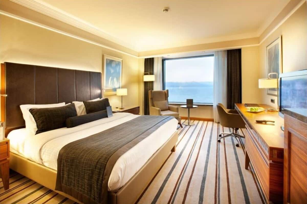 A hotel room with a large bed, a desk, a chair, a TV, and a view of the sea.