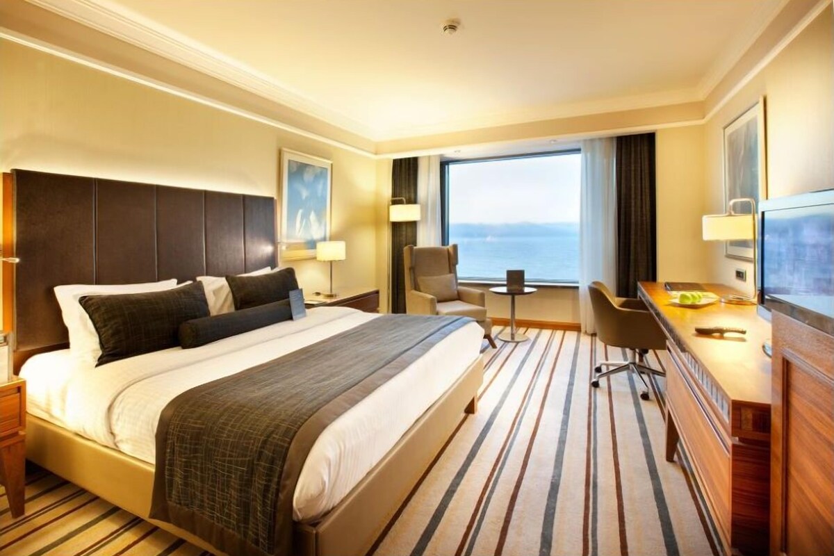 A hotel room with a large bed, a desk, a chair, a TV, and a view of the sea.
