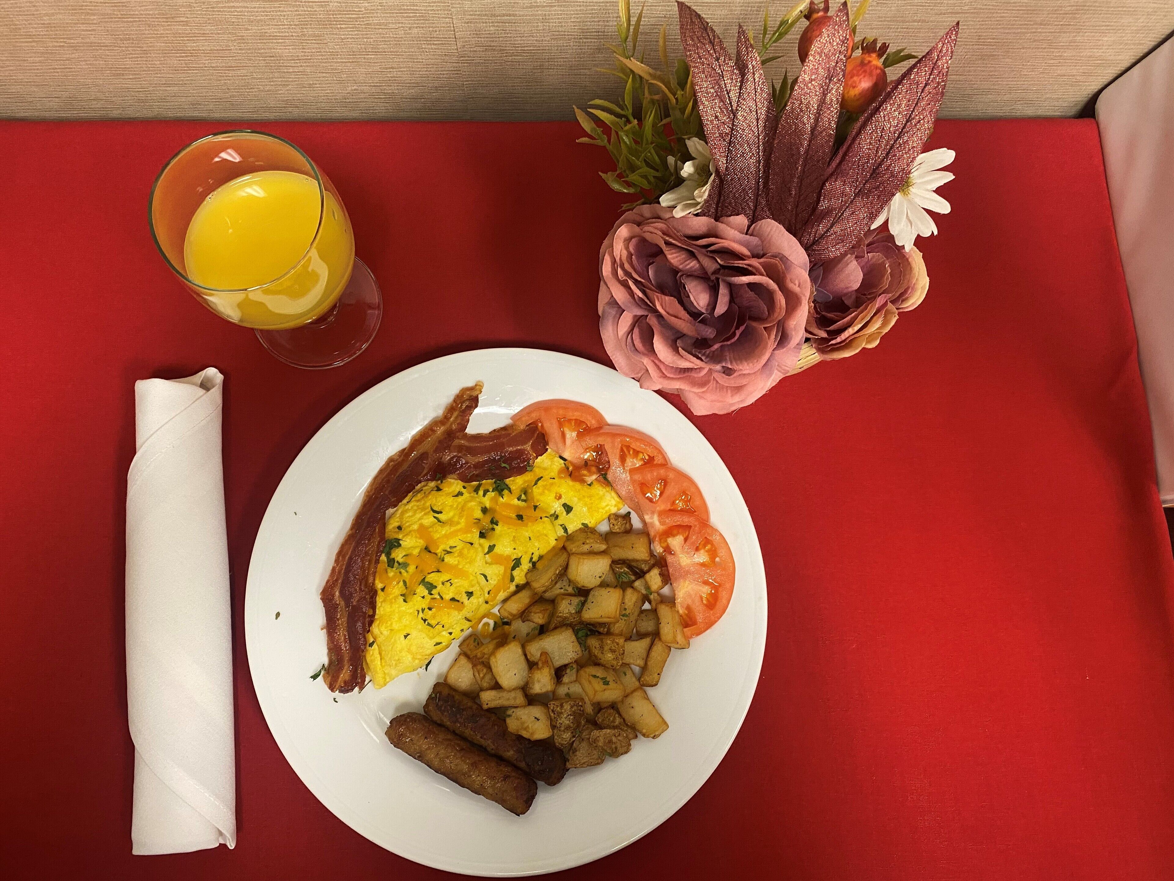 Une assiette de petit-déjeuner avec des œufs, du bacon, des pommes de terre et des saucisses, un verre de jus d’orange et une serviette sur une nappe rouge.