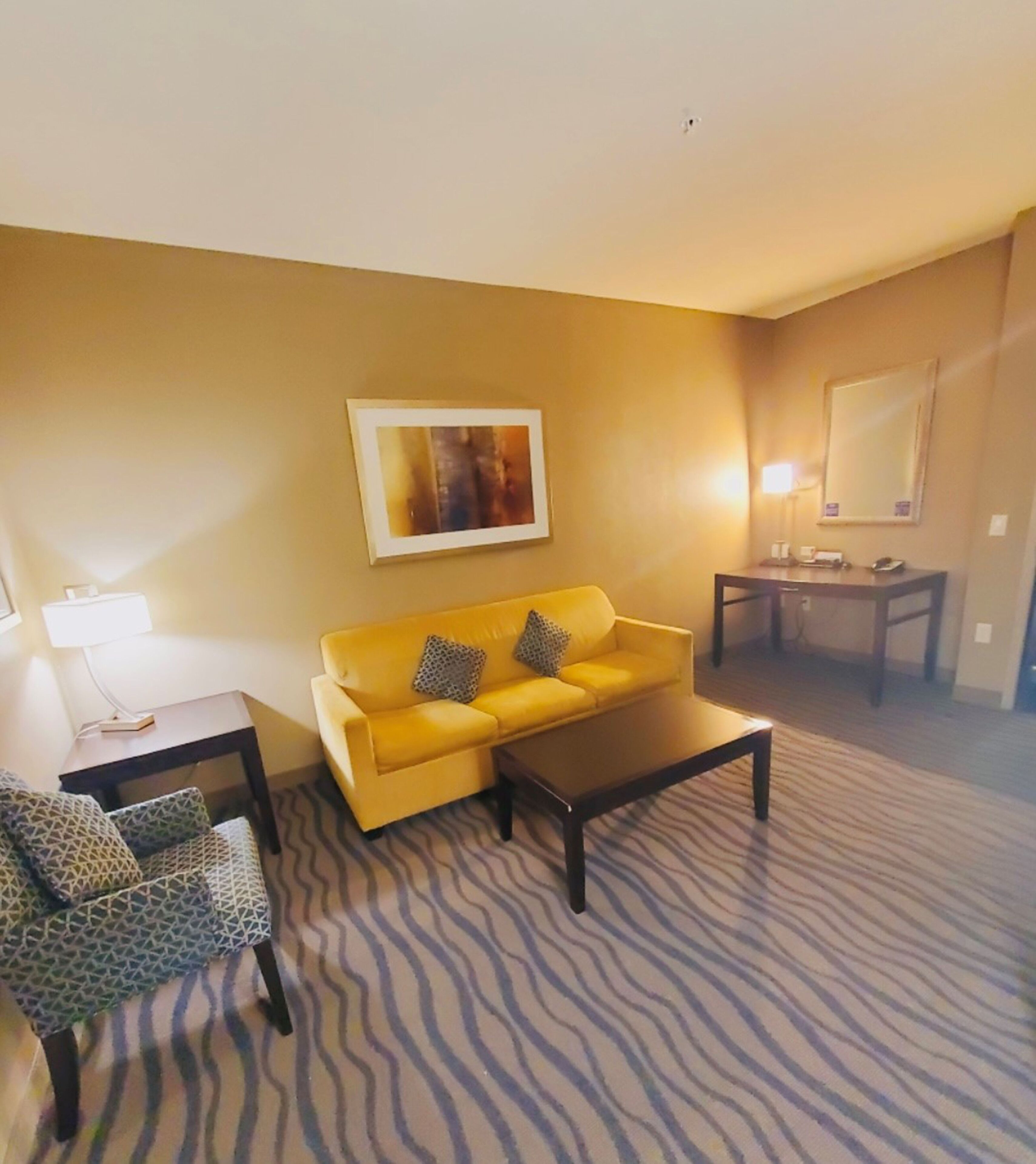 A living room with a yellow sofa, patterned armchair, and a coffee table. There is a painting on the wall and a lamp on a side table.