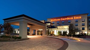 Exterior de un Hilton Garden Inn con entrada cubierta, fachada bien iluminada y zona ajardinada al frente.