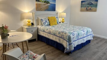 Um quarto com uma cama, duas luminárias de cabeceira, uma mesinha de cabeceira, uma mesa redonda com uma cadeira e um quadro na parede.