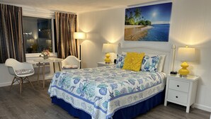 A bedroom with a bed, bedside tables, a chair, a desk, and a painting of a beach scene. - Mariner Motel (Falmouth)